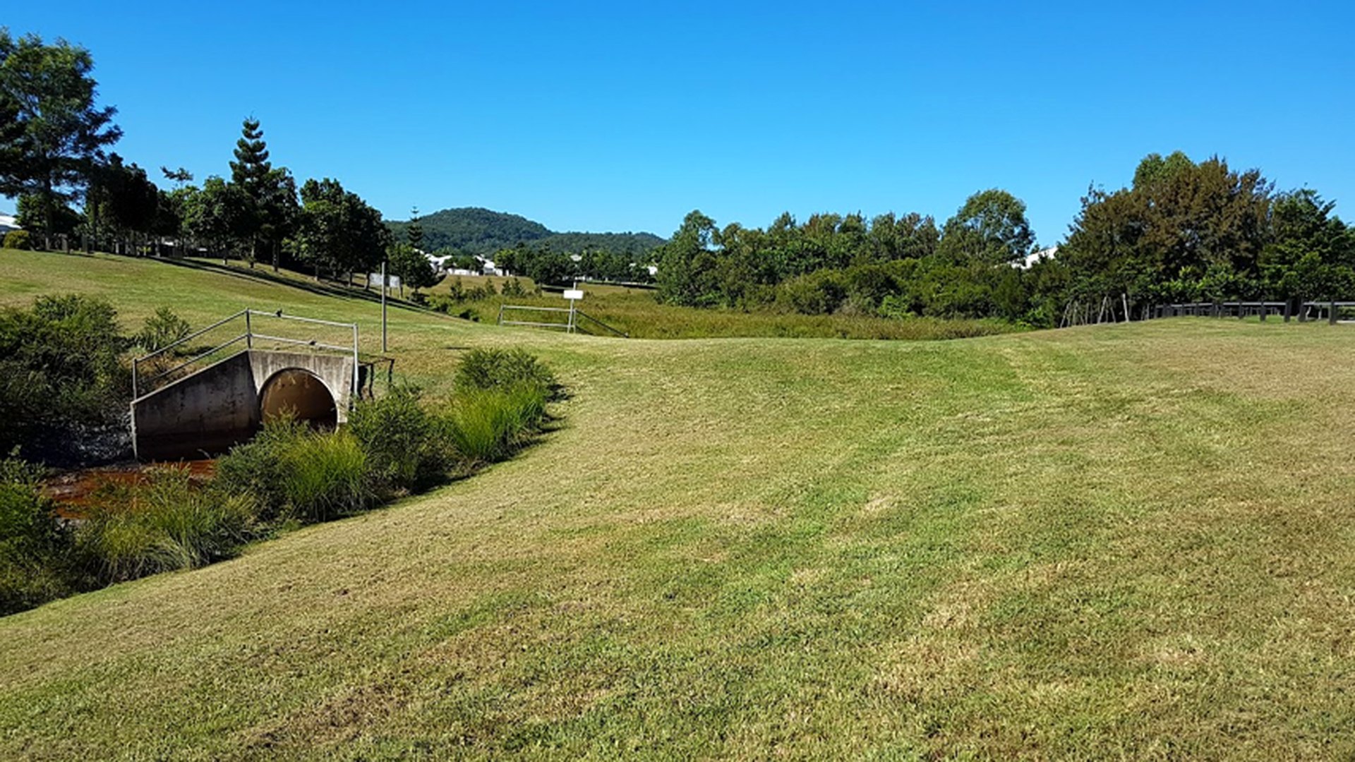 Wide mown grassed parkland and embankment beside a culvert bridge under clear blue sky